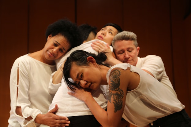 Five performers embrace in a huddle, their faces displaying a range of emotions from care to comfort to concern. They are all wearing white shirts.
