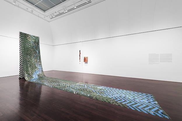 A weaved long carpet like structure of blue and green colors hangs from a makeshift wall in a room on the top and trails on the floor midway.  