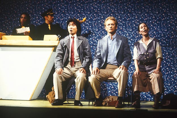Three figures in suits sit aligned with their faces a combination of surprise, wonder and singing. Behind them on the left two figures dressed as airport staff hold a yellow cable phone and tickets.  