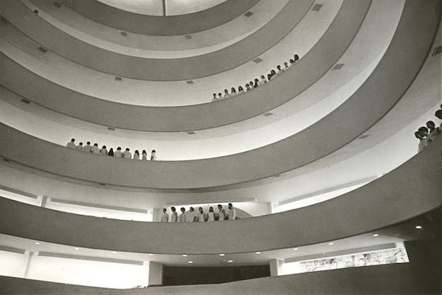 Grayscale photograph of aligned people in white standing near the balconies of different levels that meet in a swirling motion. 