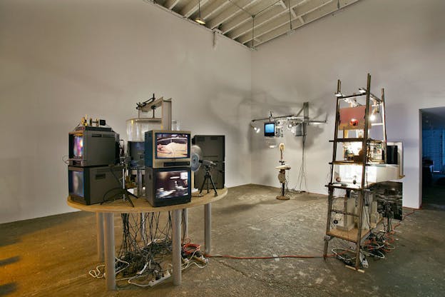 Several sculptures are situated within an otherwise bare white room. On a circle table in the foreground, there are several television screens. One shows a woman and the other shows a blurred black and white scene. Next to this table, there is a ladder with more television screens. In the background, there is a metal structure projecting from the wall which supports three addition television screens. A microphone stands below this.
