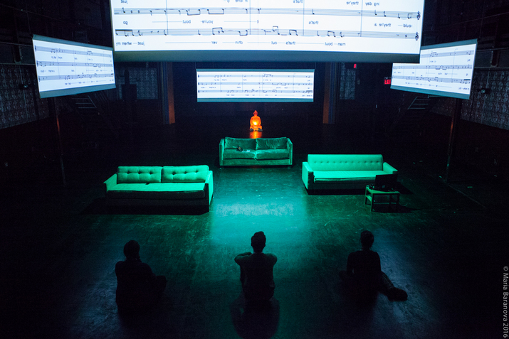 Three figures with their back towards the viewer sitting on the floor looking towards three green lighted sofas. Behind them a statue lighted orange and on top of them projections of music sheets.