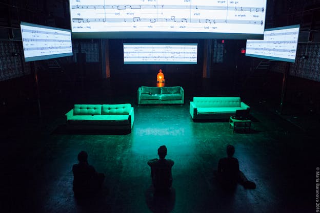 Three figures with their back towards the viewer sitting on the floor looking towards three green lighted sofas. Behind them a statue lighted orange and on top of them projections of music sheets.