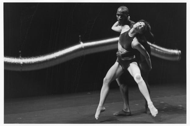 A black and white photograph of two performers in front of a long and curved aluminum duct hung from above. The first performer grabs the other performer's waist with their left hand from behind. The other performer wearing a bralette and a brief underwear embraces the neck of the first performer with their feet in the air and their eyes closed. Their head leans back onto the first performer's shoulders. The stage has a light color floor and a dark backdrop.