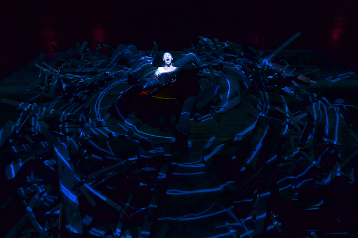 A person laying in the middle of a stage singing, with only their face lighted. surrounding them in a circular motion people laying down and peices of wood, all shadowed with only blue lines illuminating on them.