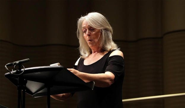 Person with gray hair shoulder-length hair and a black shirt talks behind a stand with a microphone. 