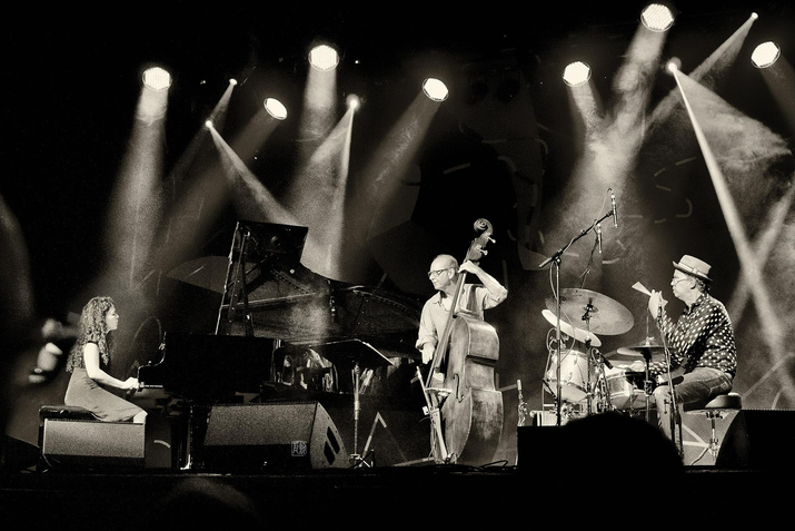 Black and white photograph of three performers on stage with multiple lights from the ceiling shining on them. On the left side a sitted figure with long curly hair plays the piano and on the righta person with a dress hat and polka dot button up plays the drums, in the middle of the two a person with glasses and a striped button up plays a body size string instrument.