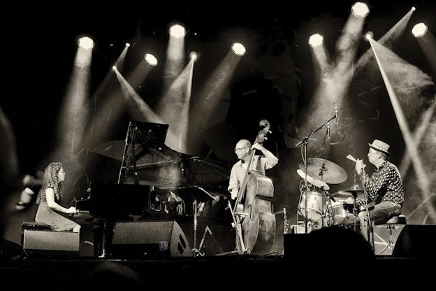 Black and white photograph of three performers on stage with multiple lights from the ceiling shining on them. On the left side a sitted figure with long curly hair plays the piano and on the righta person with a dress hat and polka dot button up plays the drums, in the middle of the two a person with glasses and a striped button up plays a body size string instrument.