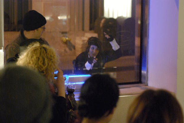 Mac Low performs, sitting behind a glass partition with a microphone in front of her. She raises her arms above her head and look intently forwards. Behind her, there are mailboxes and a fire hydrant. Audience members watch her from the opposite side of the glass partition. 