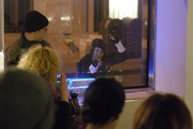 Mac Low performs, sitting behind a glass partition with a microphone in front of her. She raises her arms above her head and look intently forwards. Behind her, there are mailboxes and a fire hydrant. Audience members watch her from the opposite side of the glass partition. 