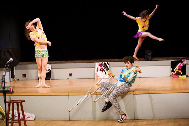 A person with a cast foot leans against the wooden stage, holding out two crutches. Behind them one performer is in mid-leap and one performer is tilting their head back and wrapping their hand over their eyes.