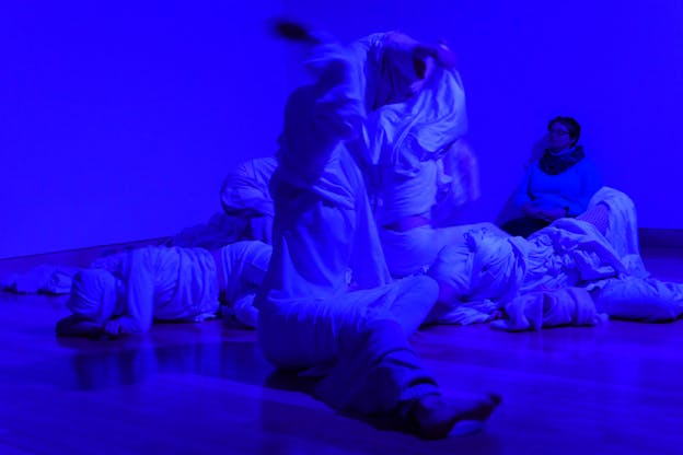 Performers wrapped in white cloths dance on the floor in a blue-lit room, their bodies blurred in motion. 