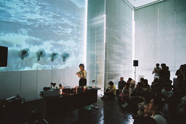 A person plays the violin behind a desk, while an audience sits on the ground crosslegged watching. Waves are projected on the background wall. 