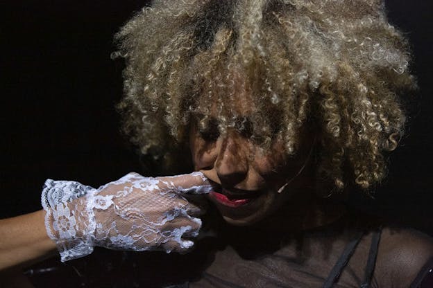 A figure with blond afro hair and red lipstick stands still as a hand in a white lacy glove inserts a finger through the side of their mouth.