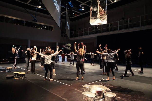 A group of people form a circle and lift their arms, standing underneath two projection screens. 