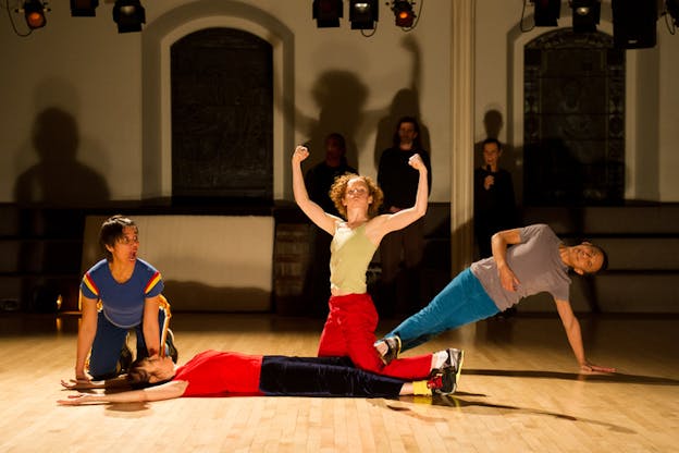 Four performers strike various poses: one lying supine, another on their hands and knees their mouth agape, one leaning on one arm, and another kneeling and raising their fists triumphantly over their head. 