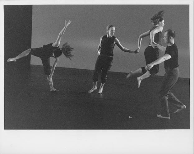 Several performers dance in a dark space. On the far left, a performer leans over one foot, throwing their arms up in the process. On the far right, a perfomer jumps aways from the camera while holding the arms of two other performers (one on each side). 