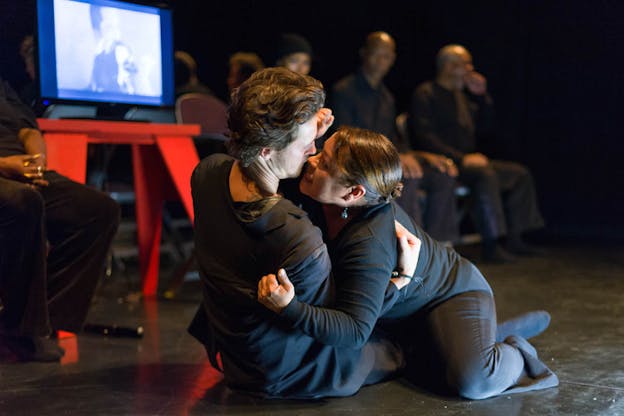 Two performers clad in black lay on the floor with one on top the other, their faces almost touching. 