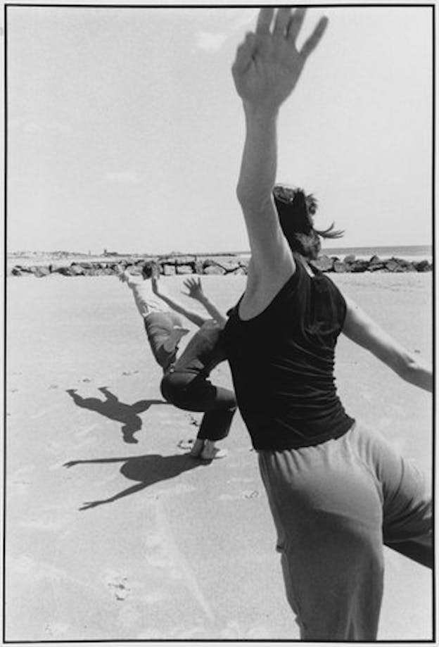 A black and white performance still of three performers in loose pants and t-shirts on a bare beach. The performers are in a line. The performer farthest away from the camera lunges forward and reaches their arm in front of them. The performer behind them squats downwards and raises their arms behind them. The performer closest to the camera leans forward and flings their arms behind them.
