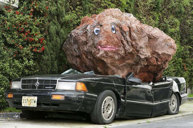 A red boulder with a face painted it on it crashed down on the roof of a cypress green car.