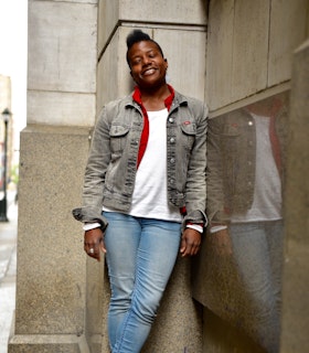 Jennifer Nikki Kidwell stands outside with her eyes closed, slightly leaning against a building’s wall and smiling. Sidewalk and streetlamps are visible on the left, out of focus. Jennifer wears jeans and a grey denim jacket over a white long-sleeved shirt and a red button-down shirt.