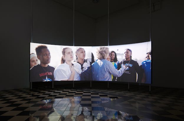 A dark gallery space with checkered stone floors and a large, concave projection screen curved to fit the shape of the room. On the projection screen, two video frames are displayed. On the left half of the screen is an image of four women athletes turned to listen to an offscreen speaker. One is wearing a crewneck that says, 