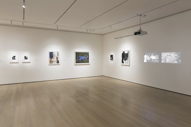 A gallery room with light wooden floors and framed photographs of various sizes lining the walls. Video works are being projected next to the photographs on one wall. Many are works in black and white, some depict landscapes and others depict human subjects. 