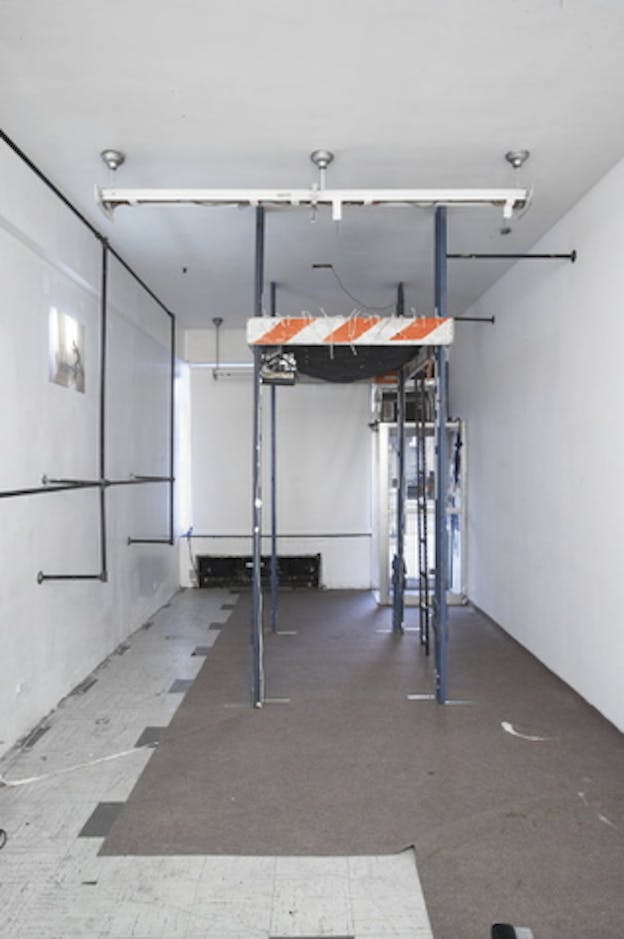 An installation image of a four floor-to-ceiling blue metal rods arranged on the corners of a square. These rods support an orange and white striped horizontal object between them. Two black metal rods connect two of the blue rods to the white wall. On another white wall, there is a rectangular structure made up of the same black metal rods. In the center of this object, on the wall, there is a small rectangular projection of a person lunging. 