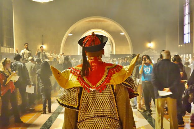 The back of a person dressed in emperor like ancient costume adorned with gold and red walks on a hall full of people.