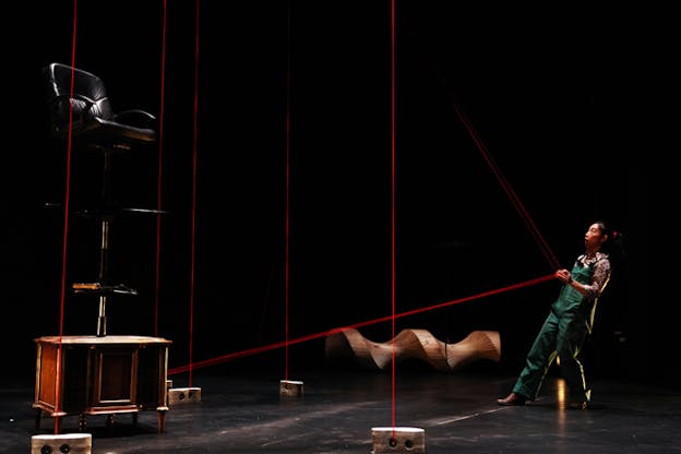 A person holding their hands together and leaning bacwards looks up as red  lines extend from their hands to various directions on the black stage. In front of them a desk support chairs balanced on one another and surrounding bricks with holes in them. 