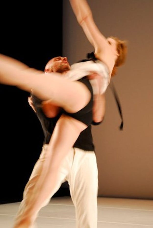 A motion shot of two performers dressed in black and white in a room with one light pink wall and one black wall. One performer catches the other who is lifted above the ground and has one arm and one leg extended outwards. 