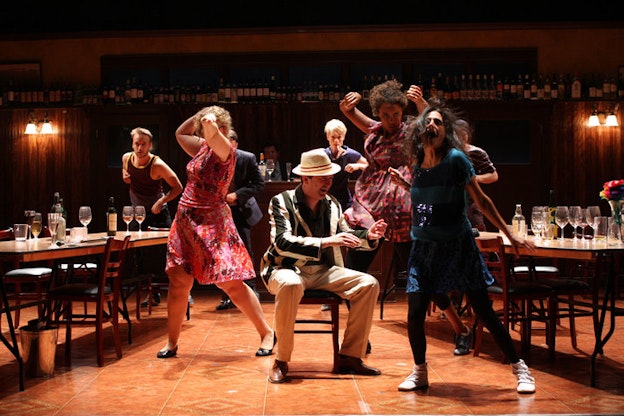 A performance still of one performer in a green and white striped suit jacket sitting on a chair in the middle of a resturant type scene. On two large table besides him, there are numerous empty wine glasses. Immediately around him, six other performers pose exuberantly with their arms thrown up behind them and their expressions intent.