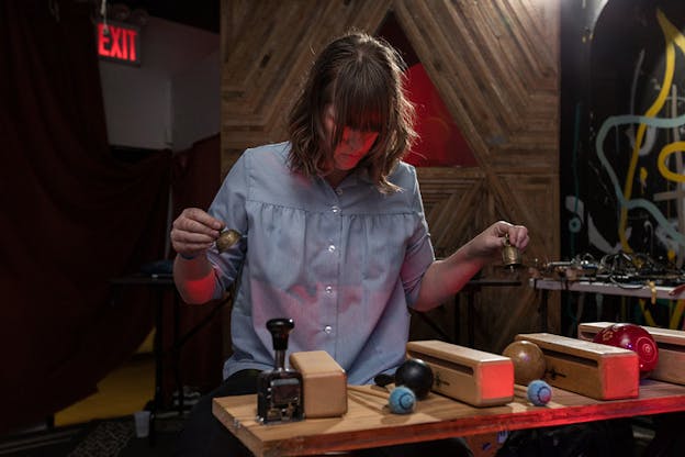 A person looking down towards a surface supporting four identical wooden slim boxes separated by various lollipop shaped instruments , holds on each hand a small bell as a red light shines on them.