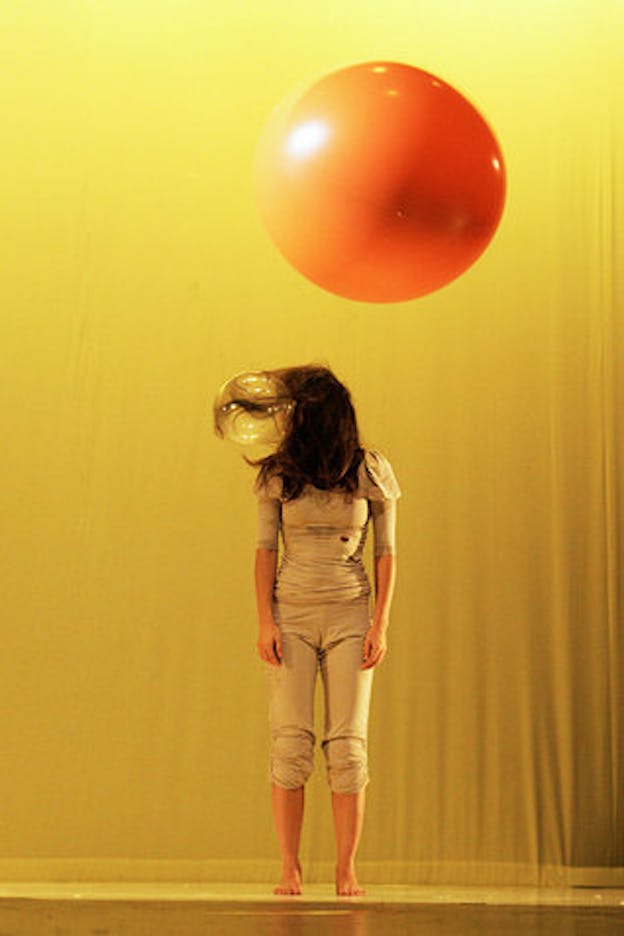 A performer wearing all beige stands in a yellow lit space. Their long brown hair covers their face and parts of a clear balloon which is suspended right next to their head. An orange sphere is suspended over them.