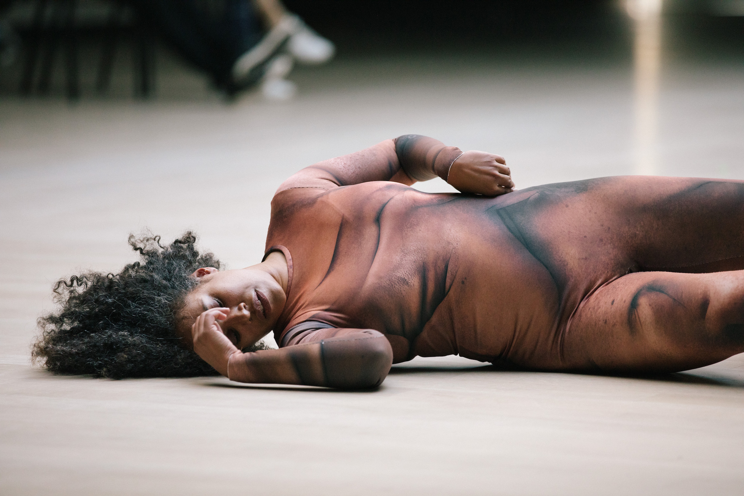 A performance still of Leslie Cuyjet laying sidewas on the ground, holding a hand to her face and closing her eyes. Her other hand rests on her hip. She wears a full length leotard printed with blurred brown and black lines. 