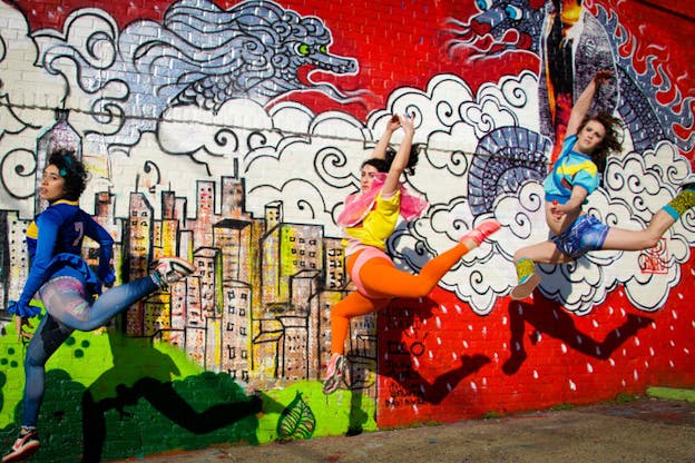 Three performers dressed in bright clothing stag leap in front of a fire-red mural depicting two dragons emerging from rain clouds above an urban skyline.