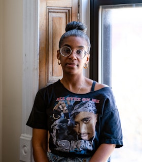 Simone White sits in a windowsill looking calmly into the camera. She wears round glasses, earrings, and an off-the-shoulder black t-shirt featuring Tupac Shakur.