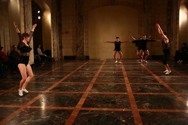 Performers clad in black leotards stand on marble floor with red square lines over it. Three of them in the back farthest from the viewer keep their arms open to their sides while another one in front of them, standing sideways raises their hands. On the left closest to the viewer stands another performer with raised arms and palms facing upwards. 