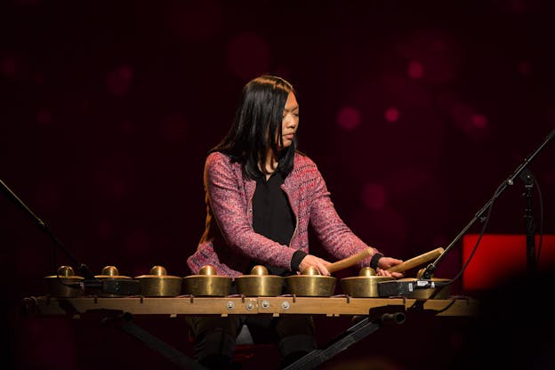 A photograph of Susie Ibarra performing with a kulintang of eight gongs, tapping two gold gongs on her left with thick wooden drumsticks. She wears a pink cardigan and a black shirt. Behind her, the scene is lit with dappled magenta light. 