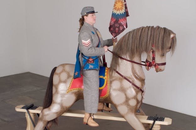 A person wearing a military uniform sits upright on a spotted horse elevated by wooden planks. The person carries a decorative quilted flag and stares resolutely ahead. 