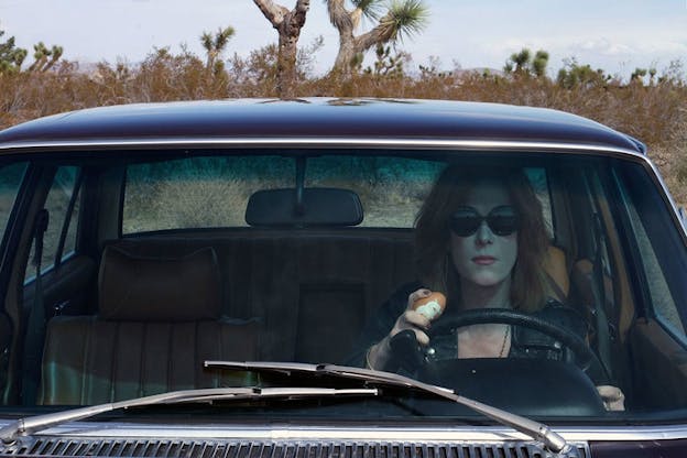 Person wearing sunglasses faces straight ahead while sitting in the drivers seat of a car parked in a desert and holding a partly-peeled hardboiled egg.