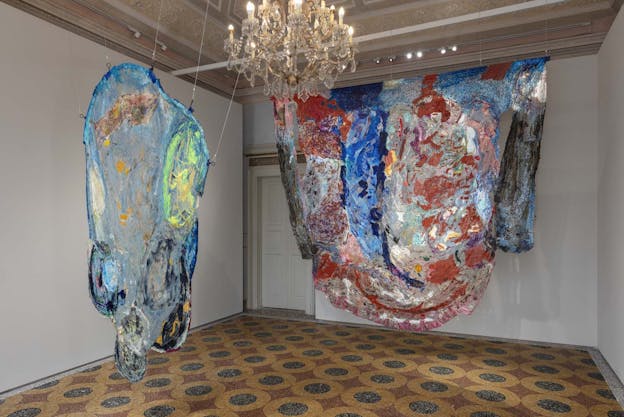 A gallery with a patterned floor, ornamental plastered ceiling, and a hanging crystal chandelier contains two large hanging sculptures. The sculpture on the left is painted blue plastic stretched over a round edged frame with protruding curves. There is a yellow oval lightly painted in the top right, and black painted curves in the rounded edges of the bottom of the piece. The sculpture on the right is draped from a straight, horizontal rod. Also painted material, this piece has swathes of red, dark blue, and flecks of black and white paint.