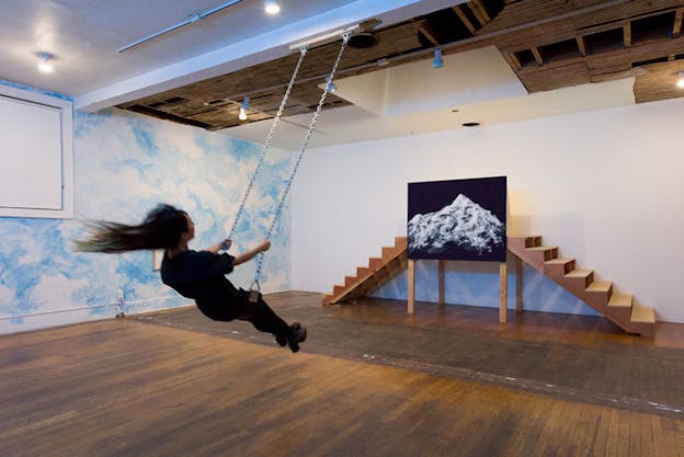 A figure dressed in black with long hair swings on a swing with metallic links hanging from the ceiling. The person faces the painting of a mountain on a black background. On either side of the painting small wooden steps that lead to its top.