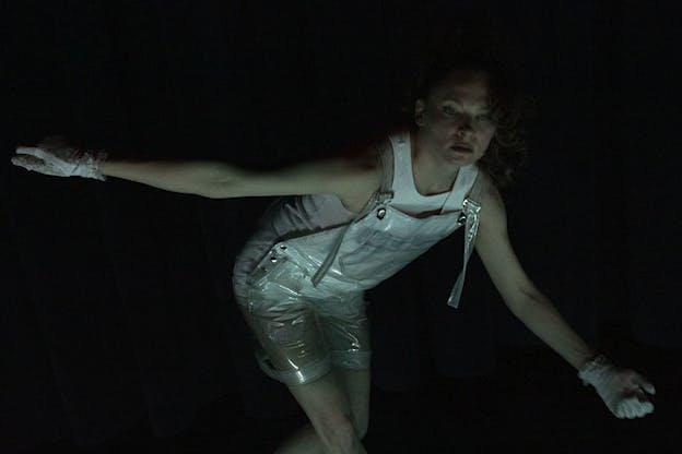 A figure in a white tank top, white shorts and transparent overalls crouches down with their arms open in a dark lit space.