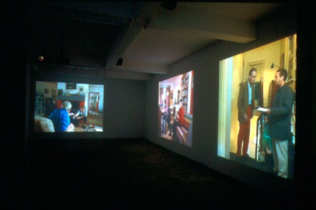Three horizontal images are projected on two walls. The image on the left is projected by itself on the wall, depicting two people sitting in a living room. The picture on the other wall being further away from the viewer appears distorted, it depicts a person playing the piano and another watching him. The third picture closest to the viewer shows a yellow background with two people talking.