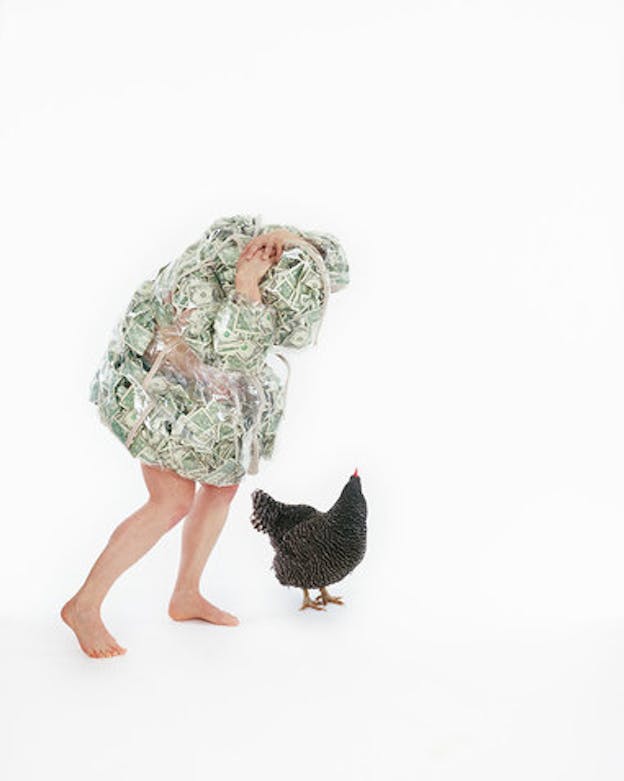 A performer wears a suit made of dollar bills and plastic which covers their head. They wrap their arms around their back and bend forward slightly. Their legs are bare. In an otherwise blank white space, there is a black chicken standing next to the performer. 
