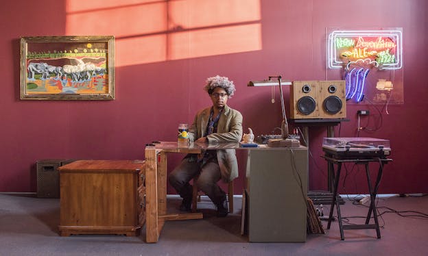 Azikiwe Mohammed sits behind a desk, against a red wall. Behind him, to the right there is a record player and a neon sign hanging on the wall that reads “New Davonhaime ALE est. 1921.” Behind him to the left is a colorful painting of elephants.