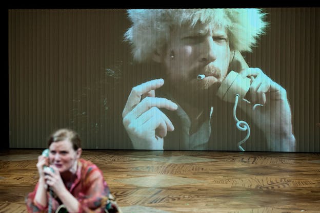 A performer's face making an emotional expression while speaking on the telephone is blurred in the front left of a stage where a man's face wearing a fur cap, smoking a cigarette, and holding a telephone to his ear is projected.
