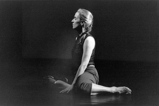 A performer sits in the middle of a dark stage with one leg bent in front of them and the other bent behind them. They look serenely in front of them at something beyond the frame of the camera.They wears a dark outfit and a braid. 