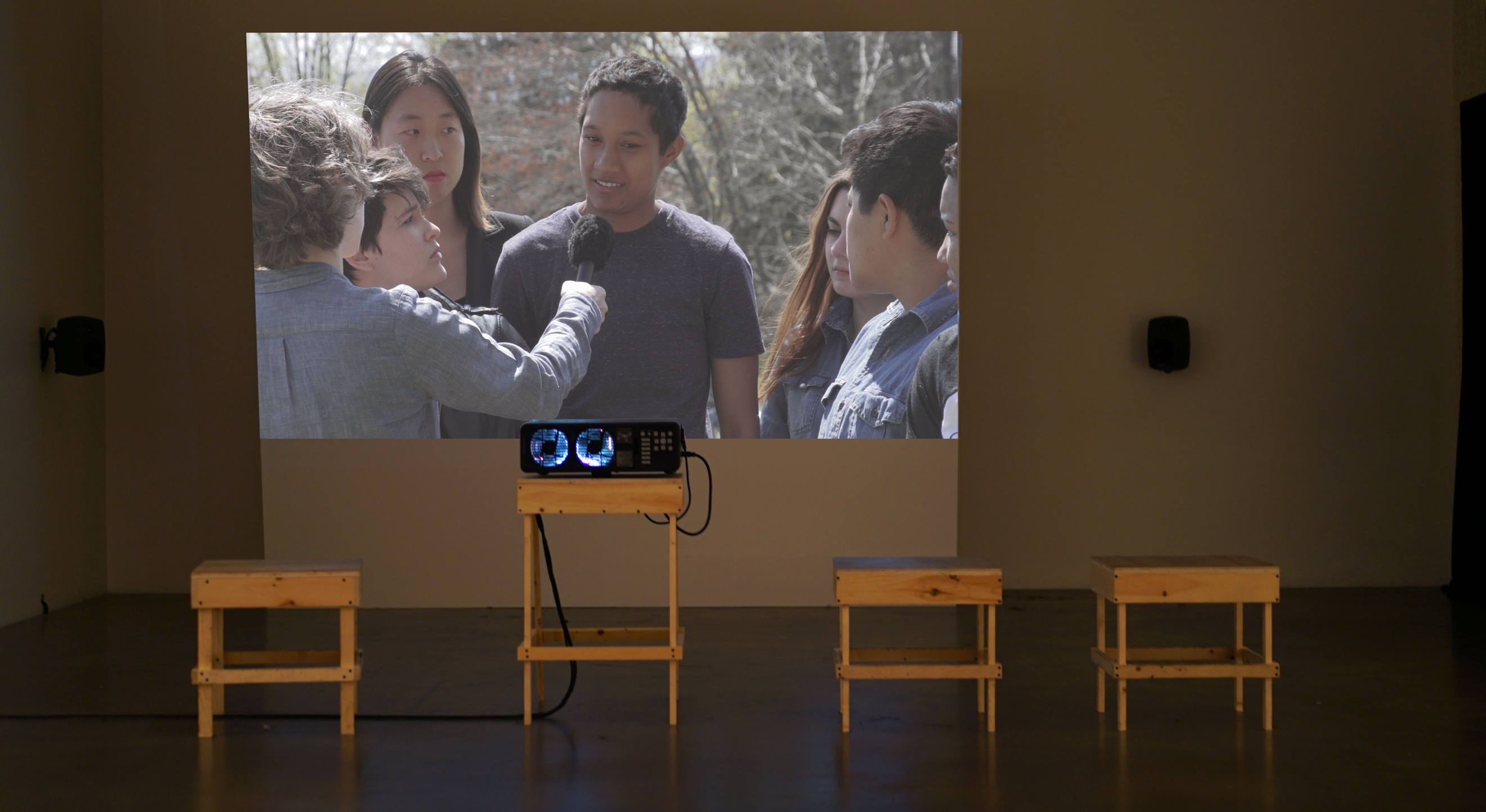 A dark gallery space with three wooden stools and a projector positioned in front of a wall with mounted speakers. In the video being projected, Sharon Hayes stands among a small group of women. She is on the left side of the frame with her back to the camera and holds a microphone up to a person across from her. 